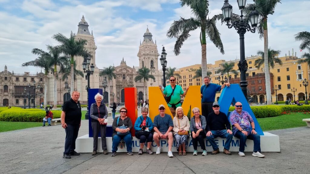 A group of senior and independent travelers posing by the giant LIMA letters in the Plaza Mayor with the Cathedral in the background during a Lima Tasty Tours historic and culinary experience.⁠