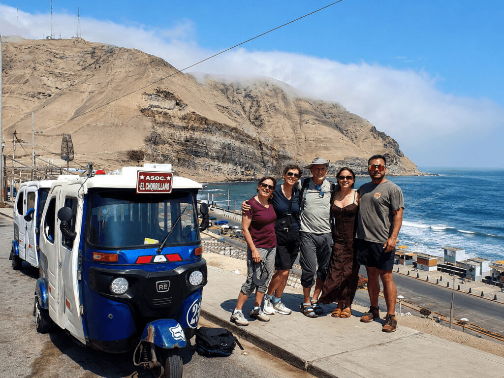 International tourists posing with a traditional blue and white Tuk-Tuk vehicle at a scenic coastal viewpoint overlooking the Pacific Ocean in Chorrillos, Lima.⁠