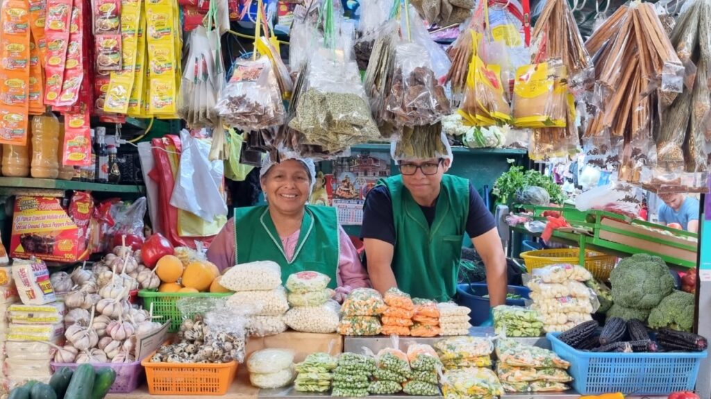Two friendly local vendors smiling behind a clean and organized traditional vegetable and spice market stall in Lima, Peru.⁠